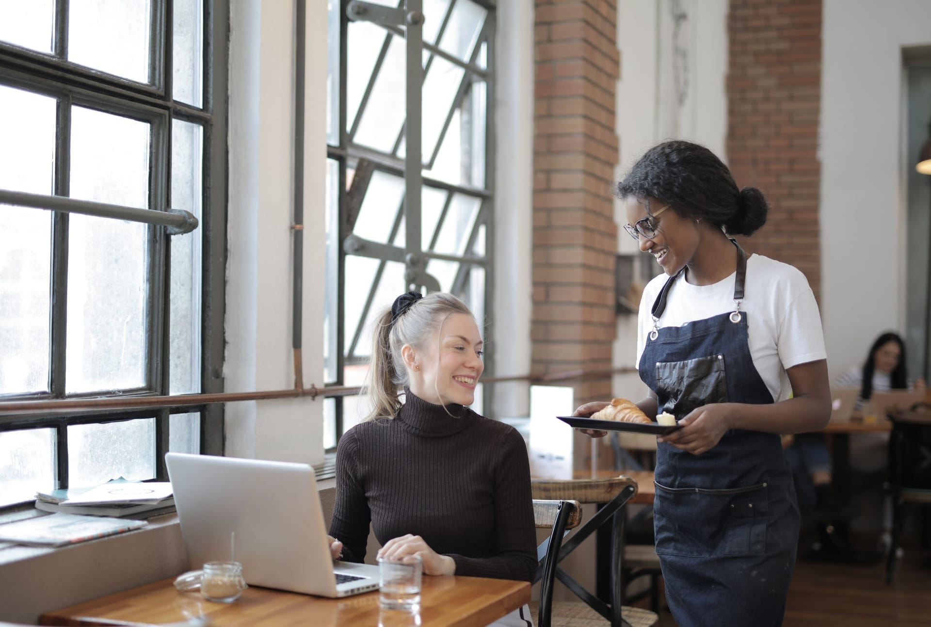 Server in an apron bringing a plate to a smiling customer at a sunlit cafe table